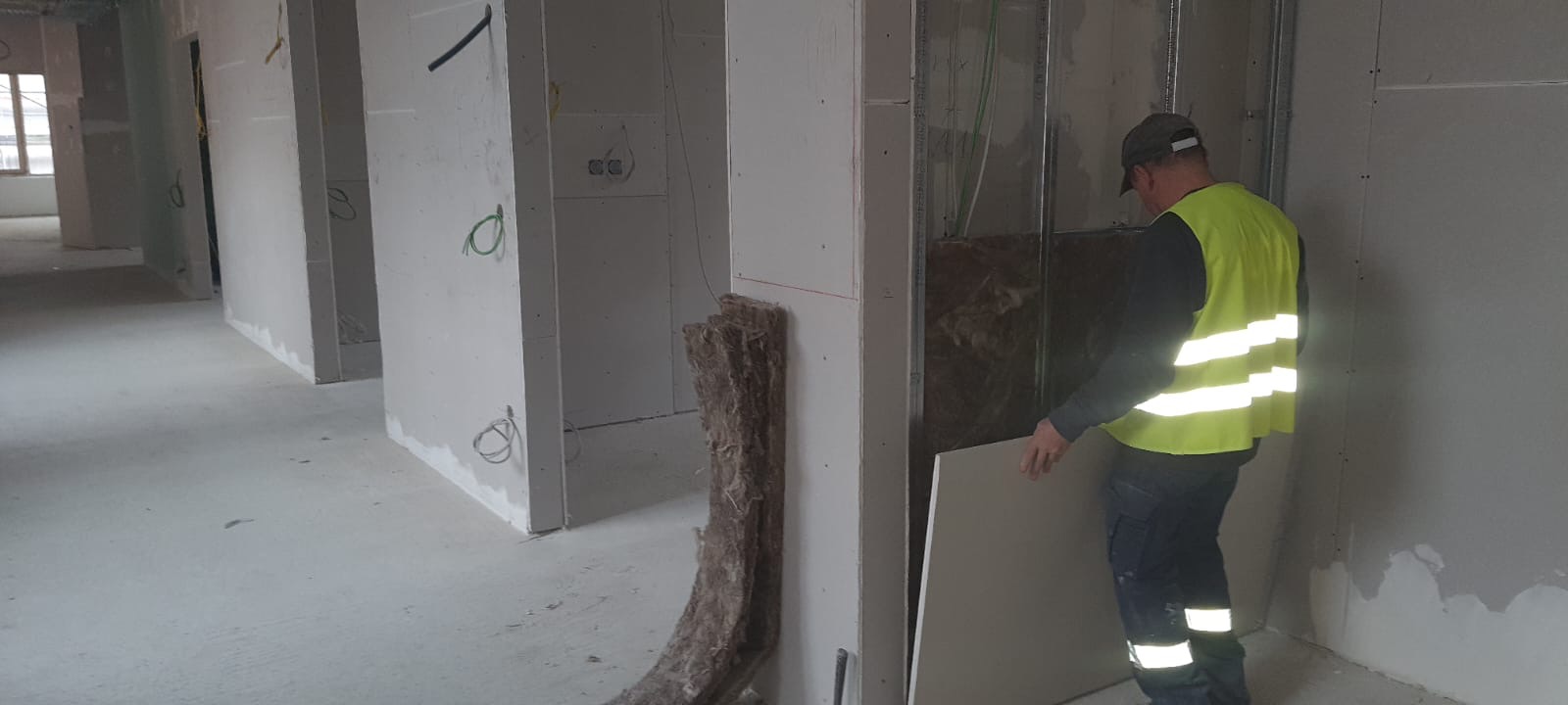 Pracownik w kamizelce odblaskowej montuje płytę gipsowo-kartonową na ścianie z izolacją z wełny mineralnej w surowym wnętrzu budynku.