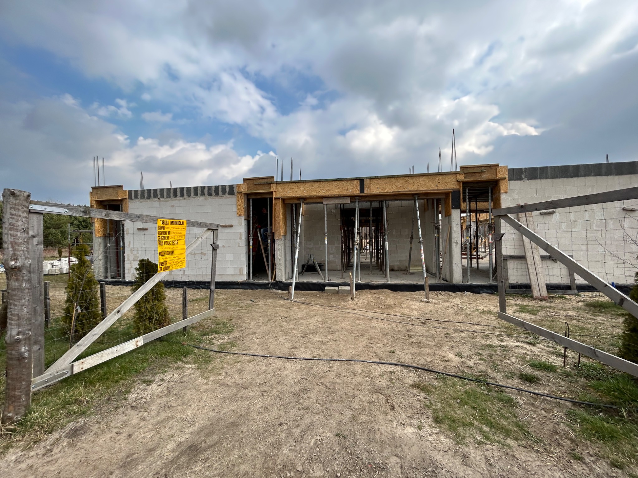 Surowy stan domu w trakcie budowy z widocznymi ścianami z bloczków, drewnianymi szalunkami okiennymi i metalowymi podporami, tablica informacyjna budowy przy prowizorycznym ogrodzeniu.