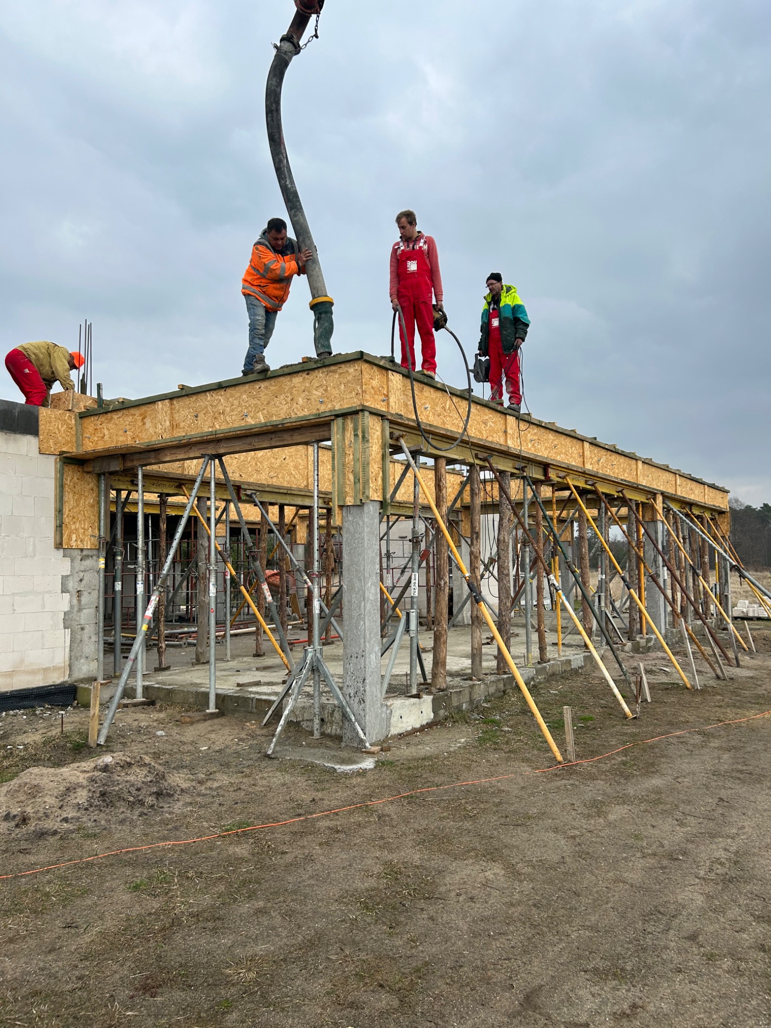 Betonowanie stropu budynku jednorodzinnego: ekipa budowlana w kombinezonach roboczych wylewa beton z pompy na szalunek z płyt OSB, wsparty stemplami i stalowymi podporami.