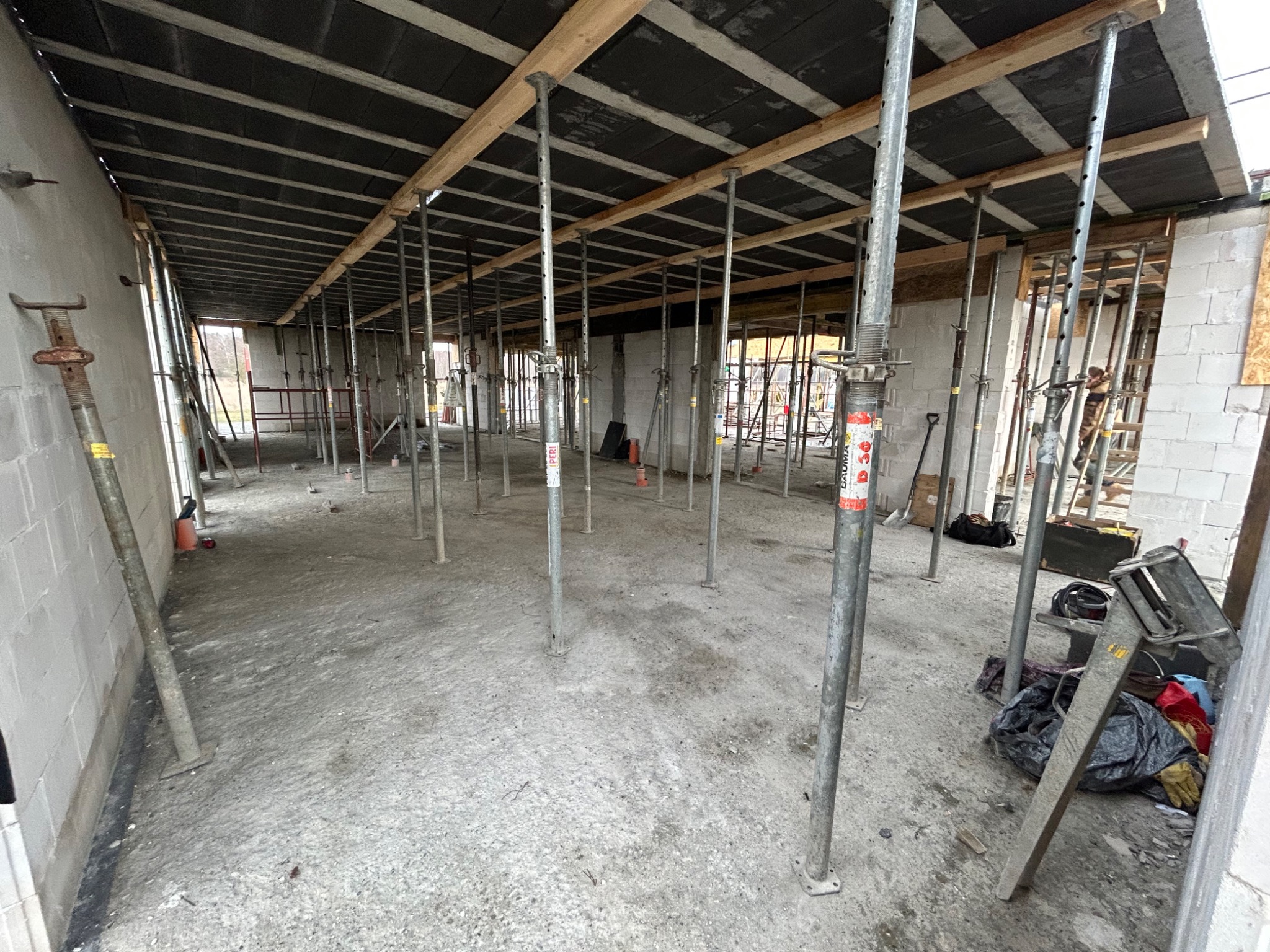 Wnętrze surowego budynku w trakcie budowy, widoczne ściany z bloczków, metalowe podpory stropu oraz drewniane belki podtrzymujące szalunek.
