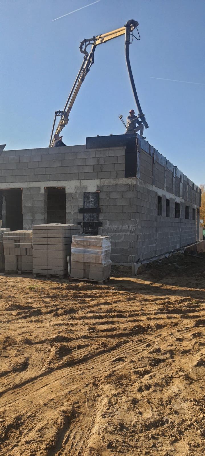 Wznoszenie murów budynku z bloczków betonowych; na górze pracownicy obsługujący pompę do betonu, w tle błękitne niebo.
