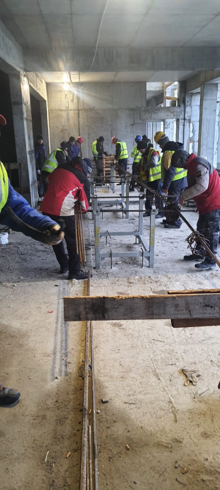 Grupa robotników w kamizelkach odblaskowych wiąże pręty zbrojeniowe na budowie, widoczne metalowe podpory i betonowe ściany w tle.