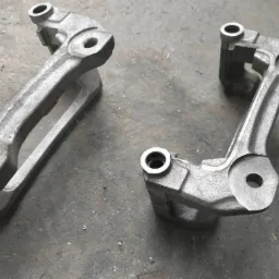 Dwa surowe, odlewane elementy metalowe o szorstkiej powierzchni, prawdopodobnie uchwyty lub wsporniki, leżące na betonowej posadzce z widocznymi drobnymi opiłkami.