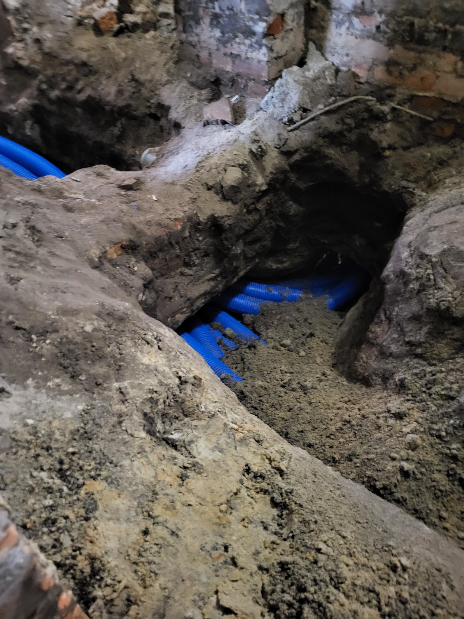 Błękitne rury karbowane ułożone w wykopie ziemnym, widoczne fragmenty ceglanego muru w tle.