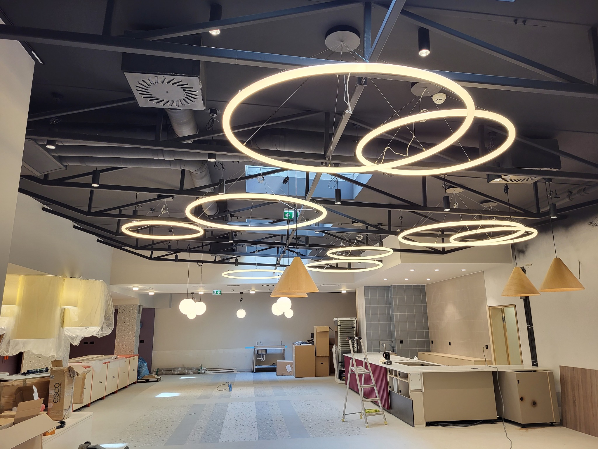 Wnętrze komercyjne w trakcie wykańczania, z widocznymi instalacjami pod sufitem, w tym okrągłymi lampami LED, wentylacją i czarnymi rurami. Na podłodze rozstawiona drabina i materiały budowlane,...