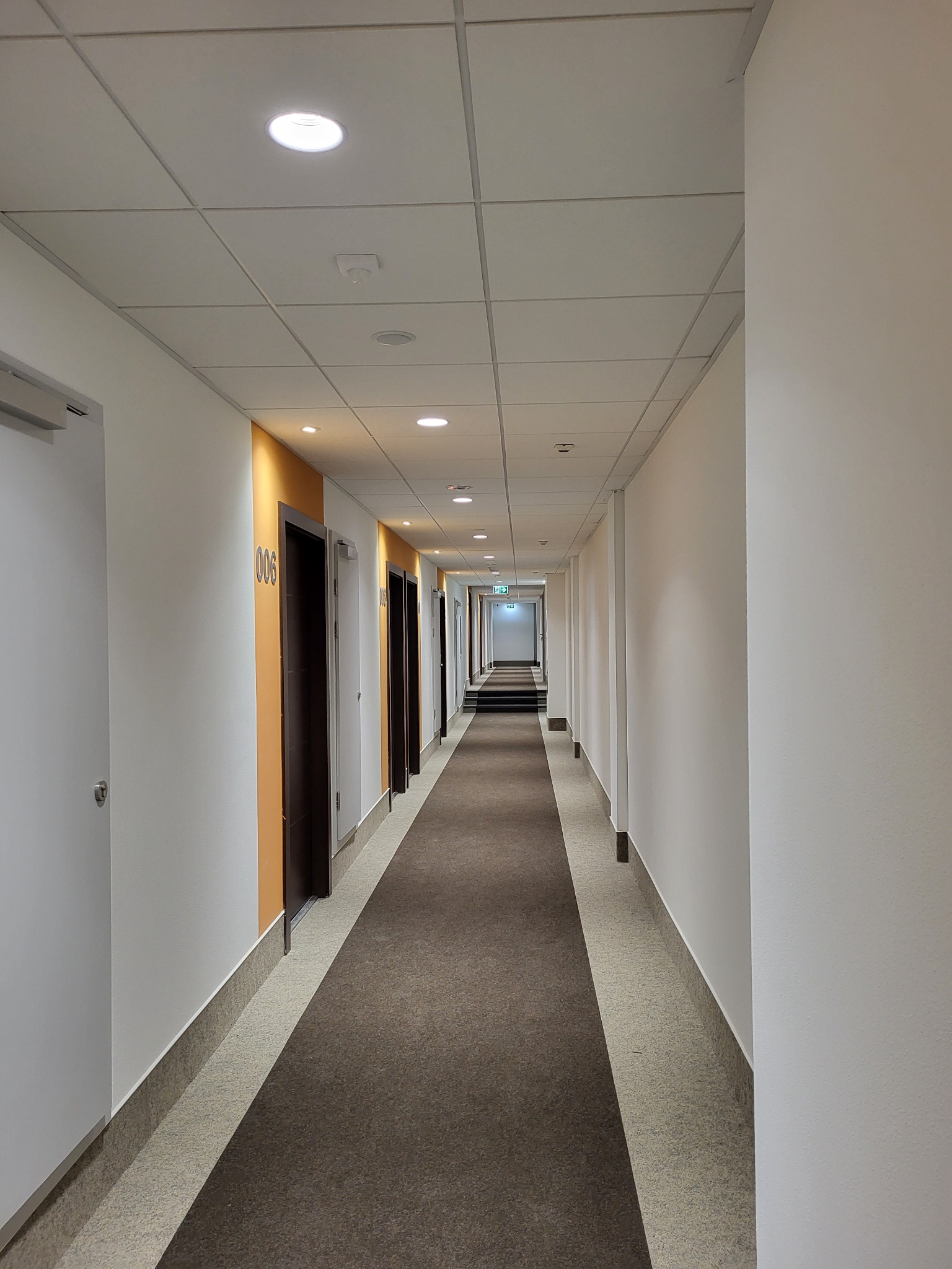 Długi korytarz biurowy z rzędem drzwi, oświetlony wpuszczanymi lampami sufitowymi. Widoczne elementy systemu przeciwpożarowego.