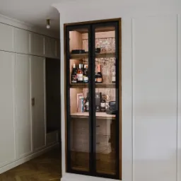 Wnęka ścienna z przeszkloną szafką na alkohol i zastawę, drewniane półki, czarne ramy, widoczny parkiet w jodełkę i fragment białej szafy w tle.
