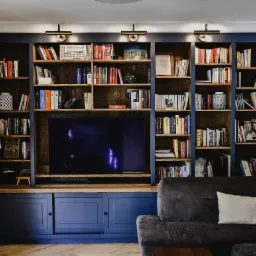 Niebieska zabudowa meblowa z regałami na książki, szafkami dolnymi i miejscem na telewizor, oświetlona lampkami, z szarą sofą w salonie.