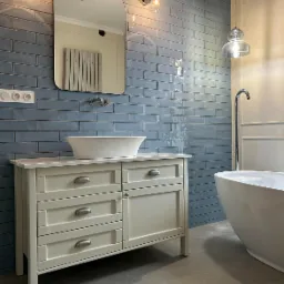 Jasna szafka łazienkowa z umywalką nablatową, na tle ściany wyłożonej niebieską, błyszczącą cegłą, z widocznym fragmentem wanny wolnostojącej i lustrem.