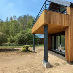 Nowoczesny dom z drewnianą elewacją i balkonem, widok z zewnątrz, teren wokół domu w trakcie zagospodarowywania, w tle las i budynek z panelami słonecznymi.