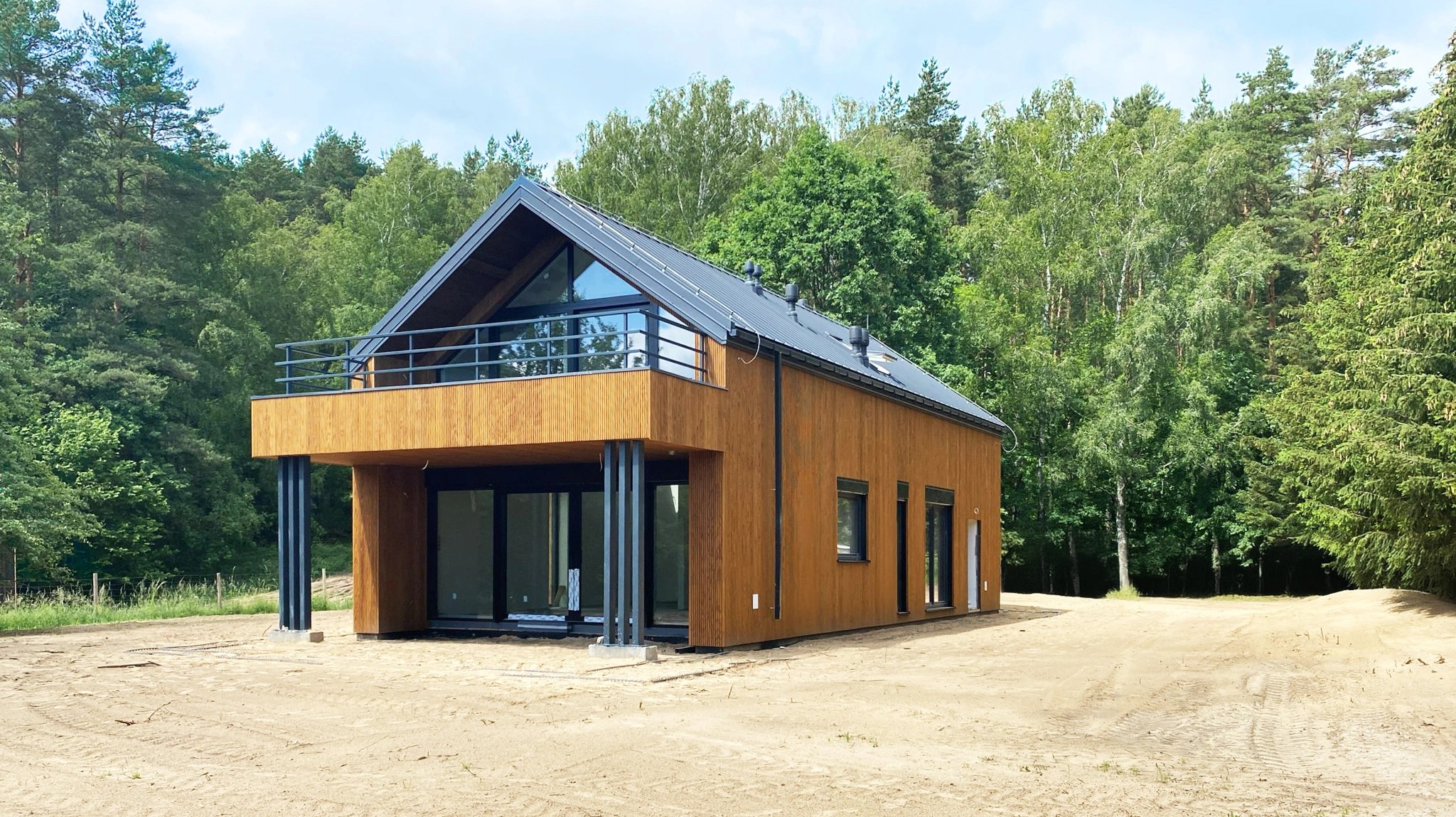 Dwukondygnacyjny dom z drewnianą elewacją, balkonem i dużymi oknami, na tle zielonego lasu, teren wokół domu przygotowany pod trawnik.