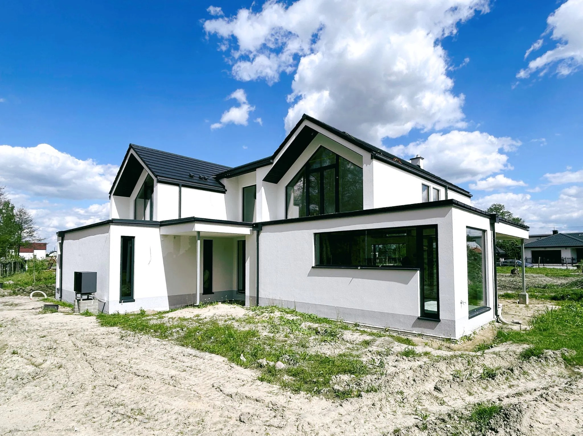 Nowoczesny, biały dom z czarnymi ramami okiennymi i dachem, widziany z zewnątrz na działce w trakcie zagospodarowywania terenu.