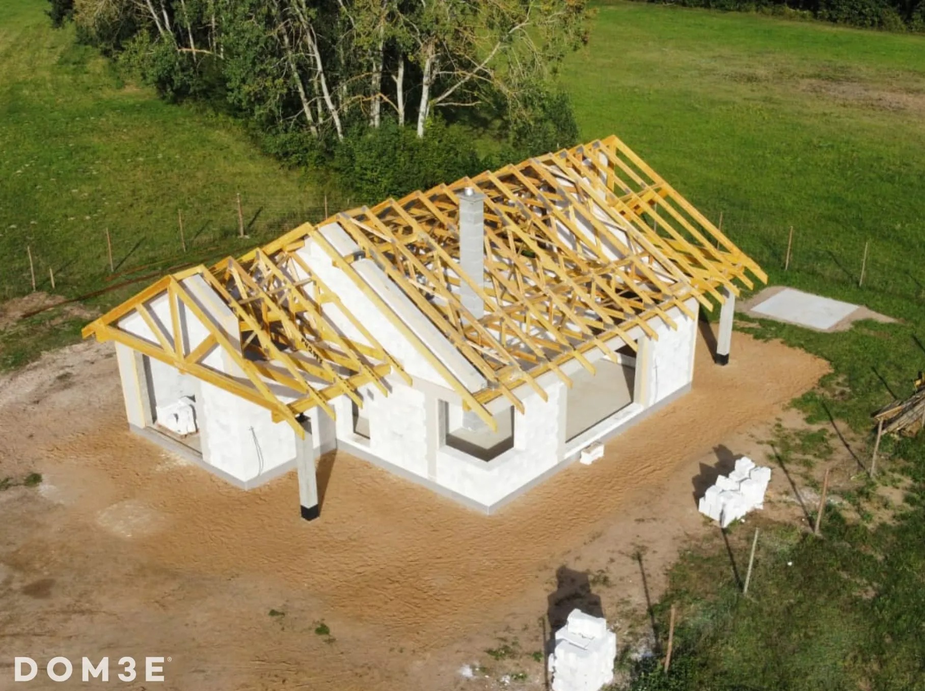 Wznoszenie domu z białych bloczków, widok z góry na konstrukcję dachu z drewnianych belek i komin, teren wokół w trakcie przygotowania.