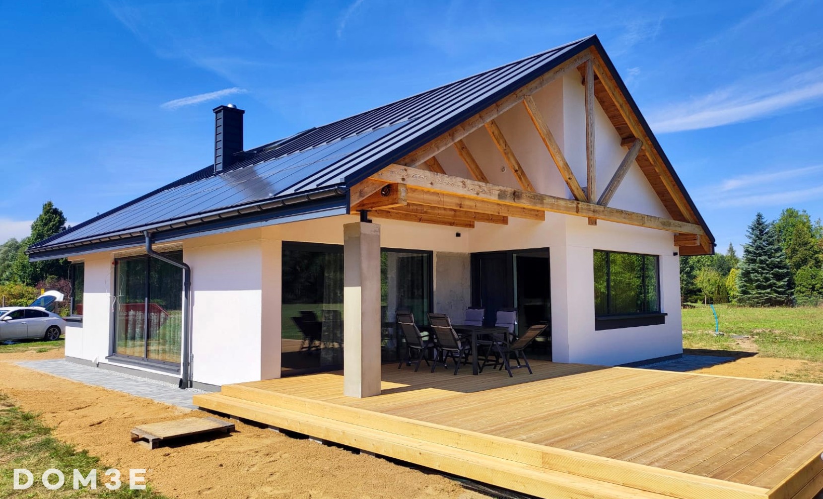 Nowoczesny dom parterowy z białymi ścianami, ciemnym dachem i drewnianym tarasem, widok z zewnątrz w słoneczny dzień.