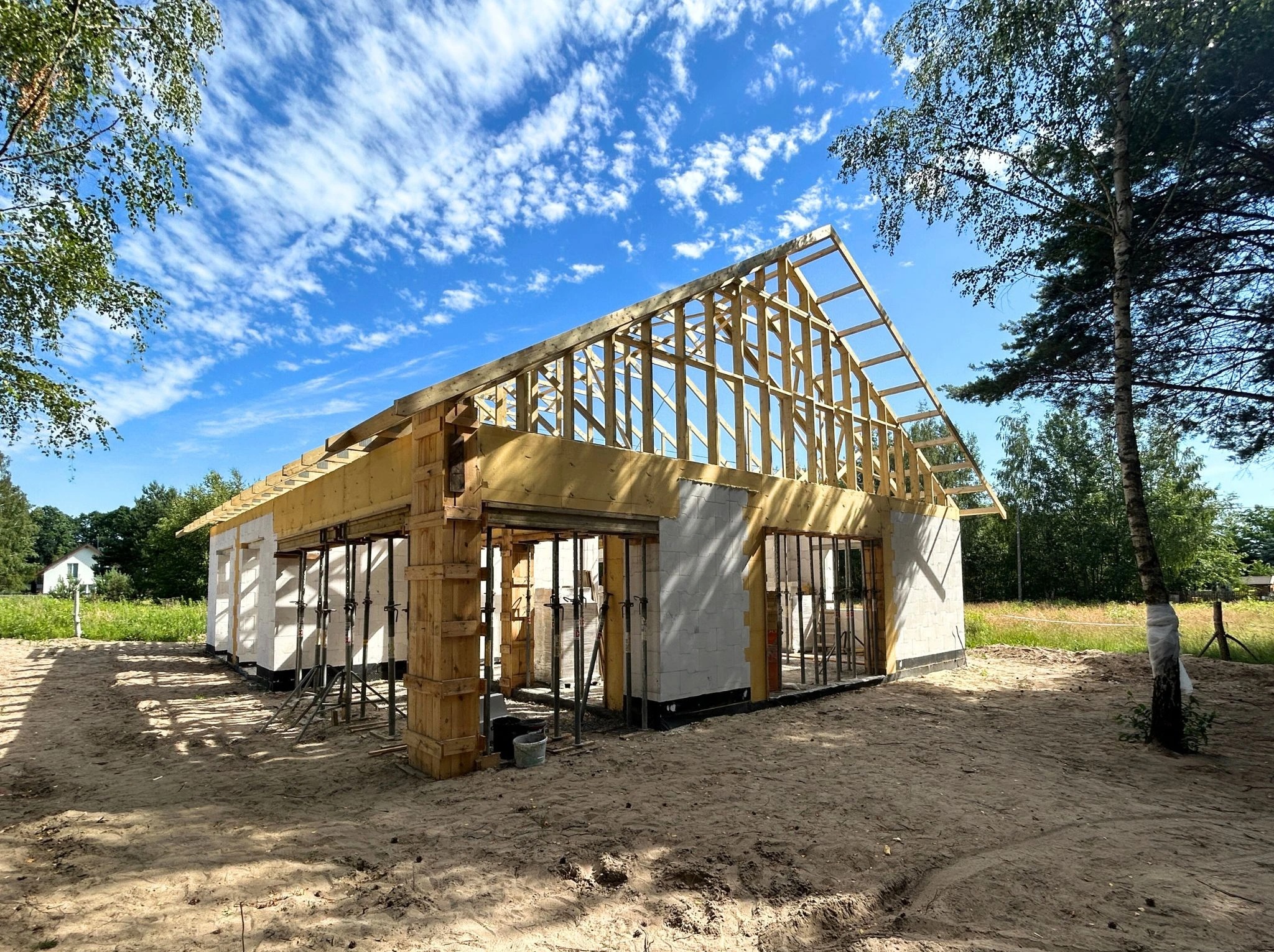 Budowa domu w stanie surowym otwartym z drewnianą konstrukcją dachu na tle błękitnego nieba z chmurami, widoczne podpory i szalunki.