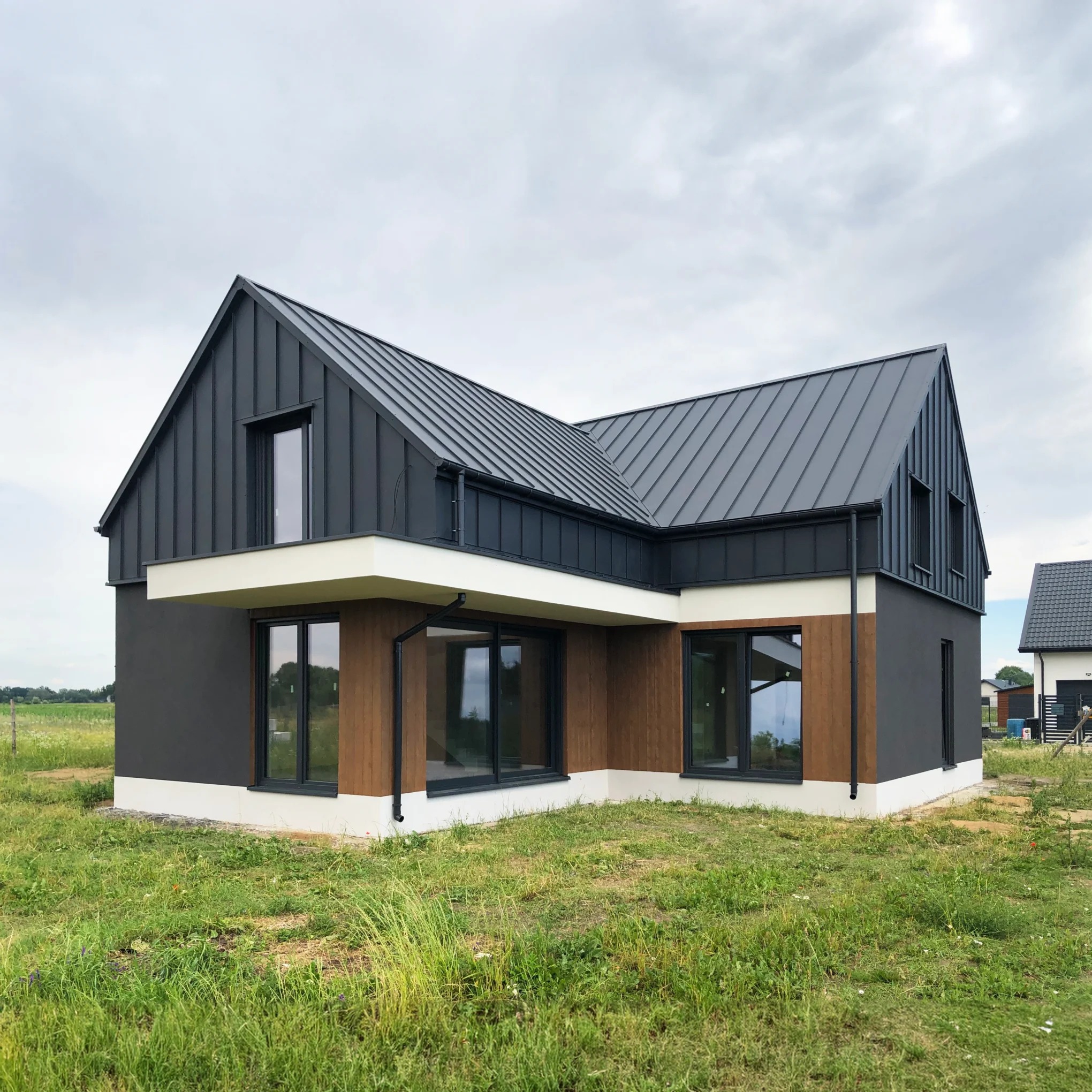 Nowoczesny dom z ciemną blachą na dachu i drewnianą elewacją w Warszawie, otoczony trawnikiem i zielenią.