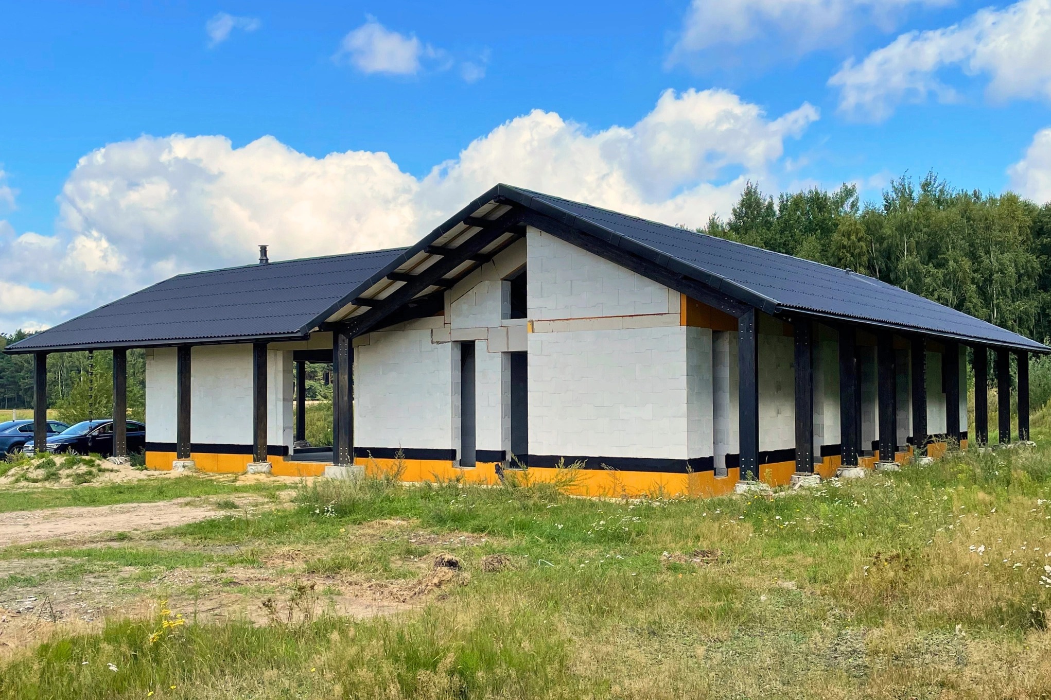 Niedokończony parterowy budynek z białych bloczków z czarnym dachem i kolumnami, widoczne żółte zabezpieczenia fundamentów, w tle las i błękitne niebo z chmurami.