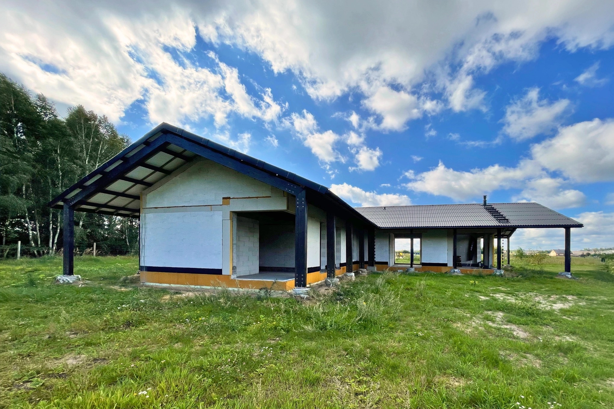 Budowa domu w stanie surowym otwartym, ściany z białych bloczków, czarny dach i elementy konstrukcyjne, widoczne z zewnątrz w otoczeniu trawy i drzew.
