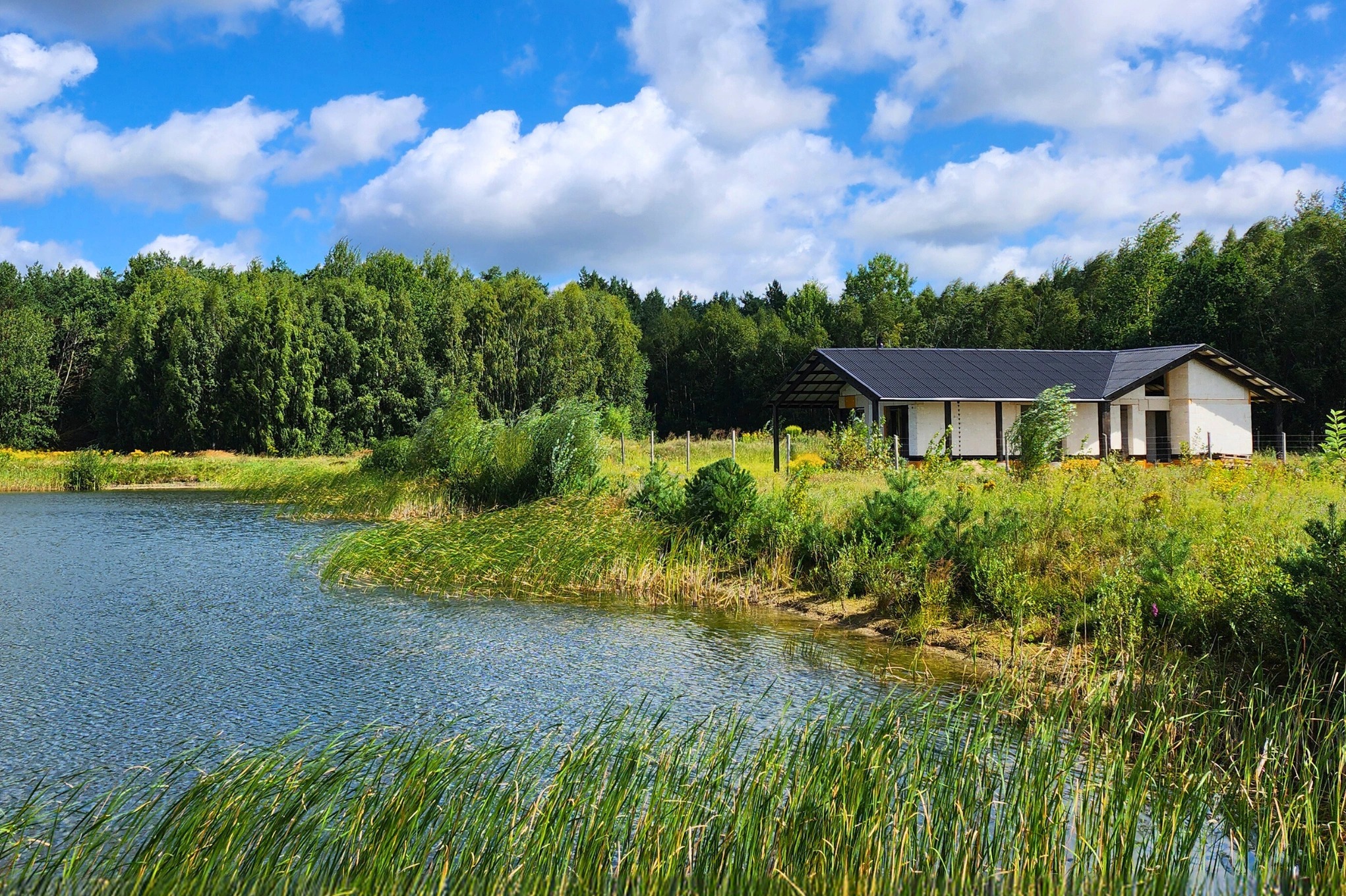 Niedokończony dom z białych pustaków, czarny dach, widok od strony jeziora z trzcinami, słoneczny dzień.