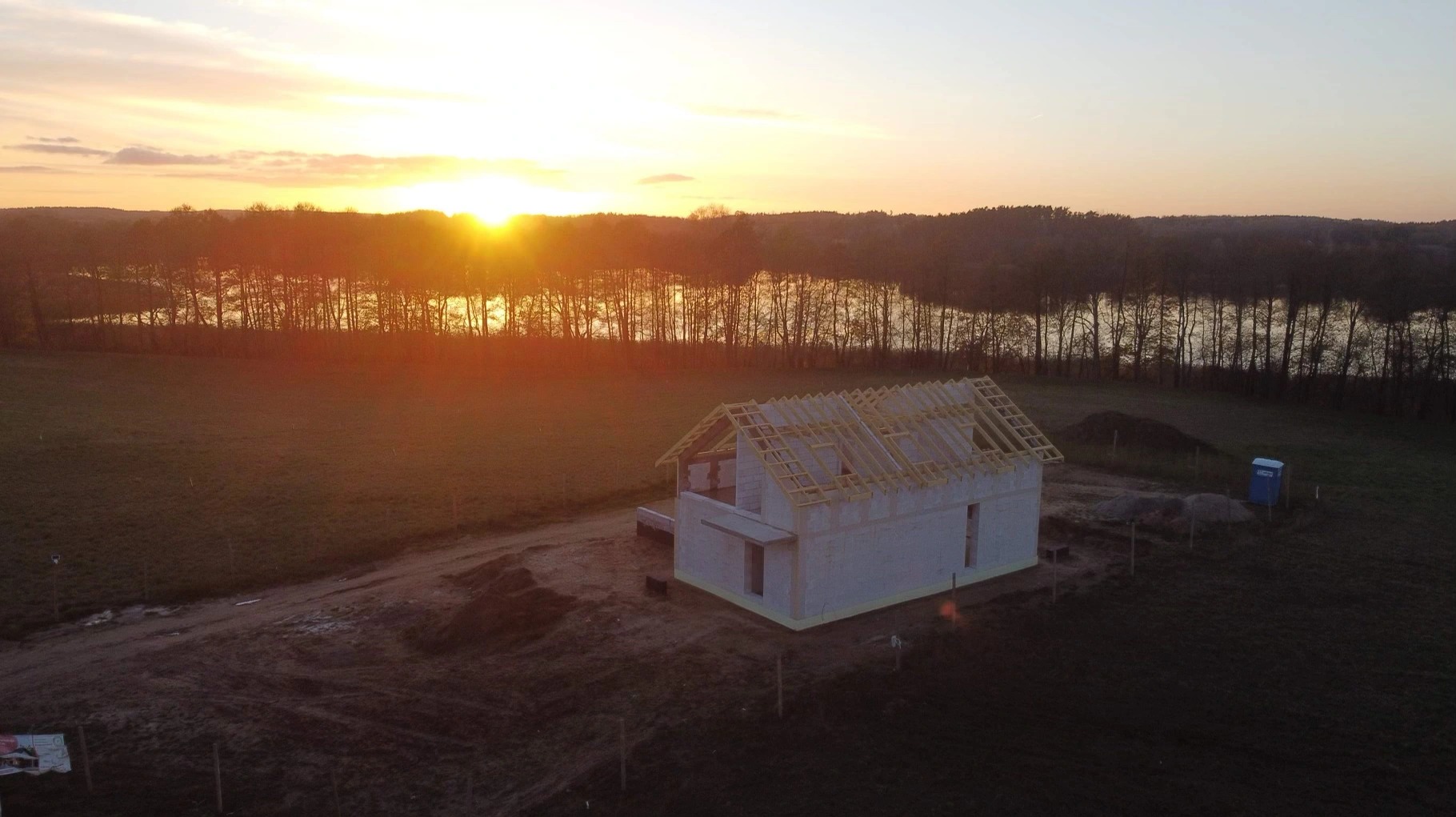 Wznoszony dom jednorodzinny z białych bloczków, widok z lotu ptaka o zmierzchu, konstrukcja dachu w trakcie montażu, w tle jezioro i zachodzące słońce.