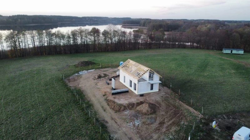 Budowa domu murowanego w stanie surowym otwartym z drewnianą konstrukcją dachu, na wzgórzu z widokiem na jezioro i las w tle, sfotografowane z lotu ptaka.