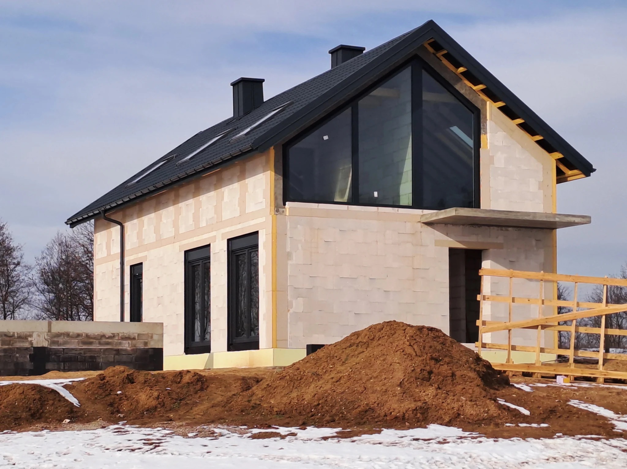 Niedokończony dom z keramzytobetonu z dużym trójkątnym oknem, czarną dachówką i oknami, na działce z resztkami śniegu i stertą ziemi.