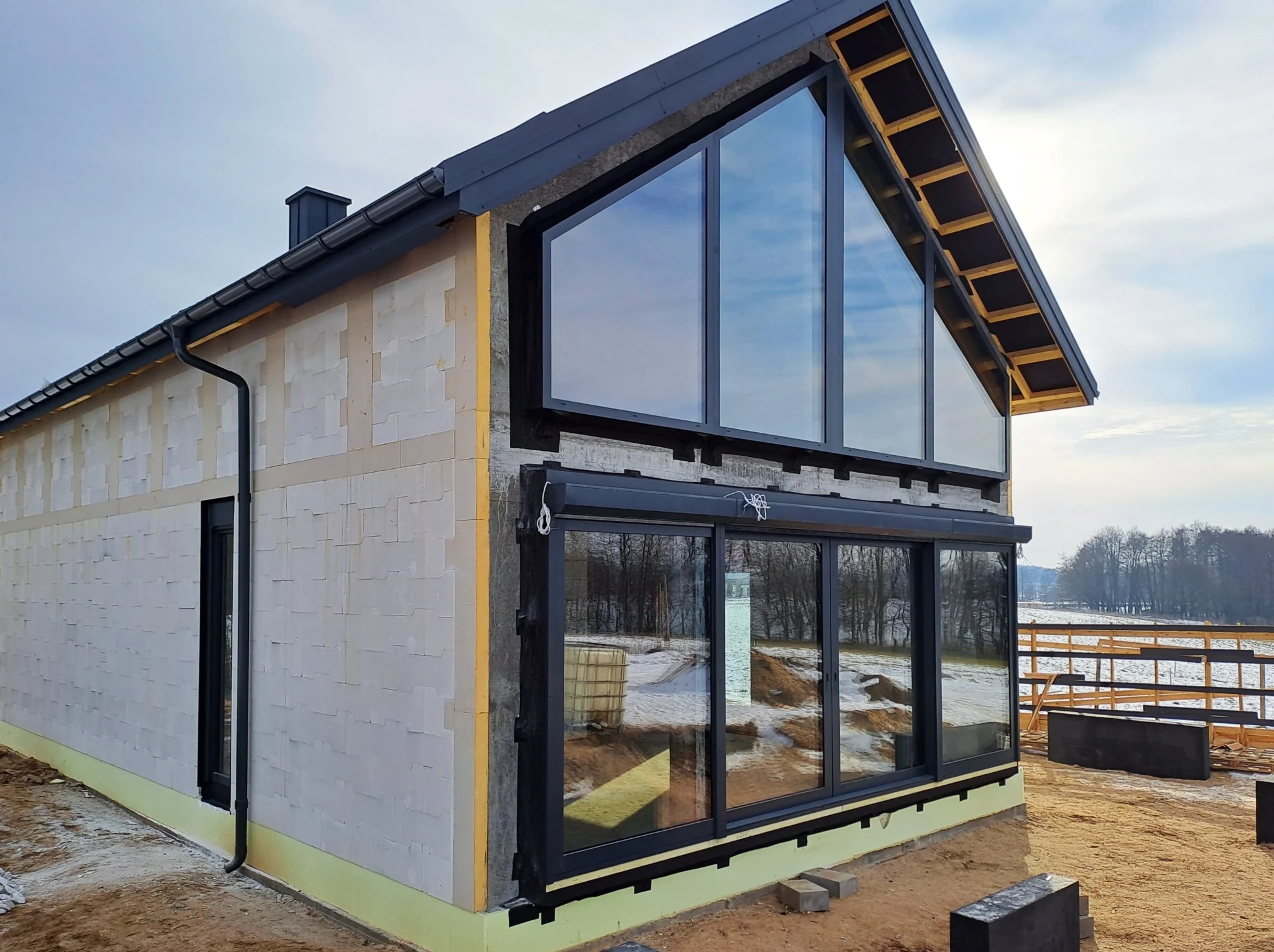 Niedokończony dom z białych bloczków z dużymi oknami, widoczne drewniane elementy konstrukcyjne dachu, teren wokół domu przygotowany pod dalsze prace.