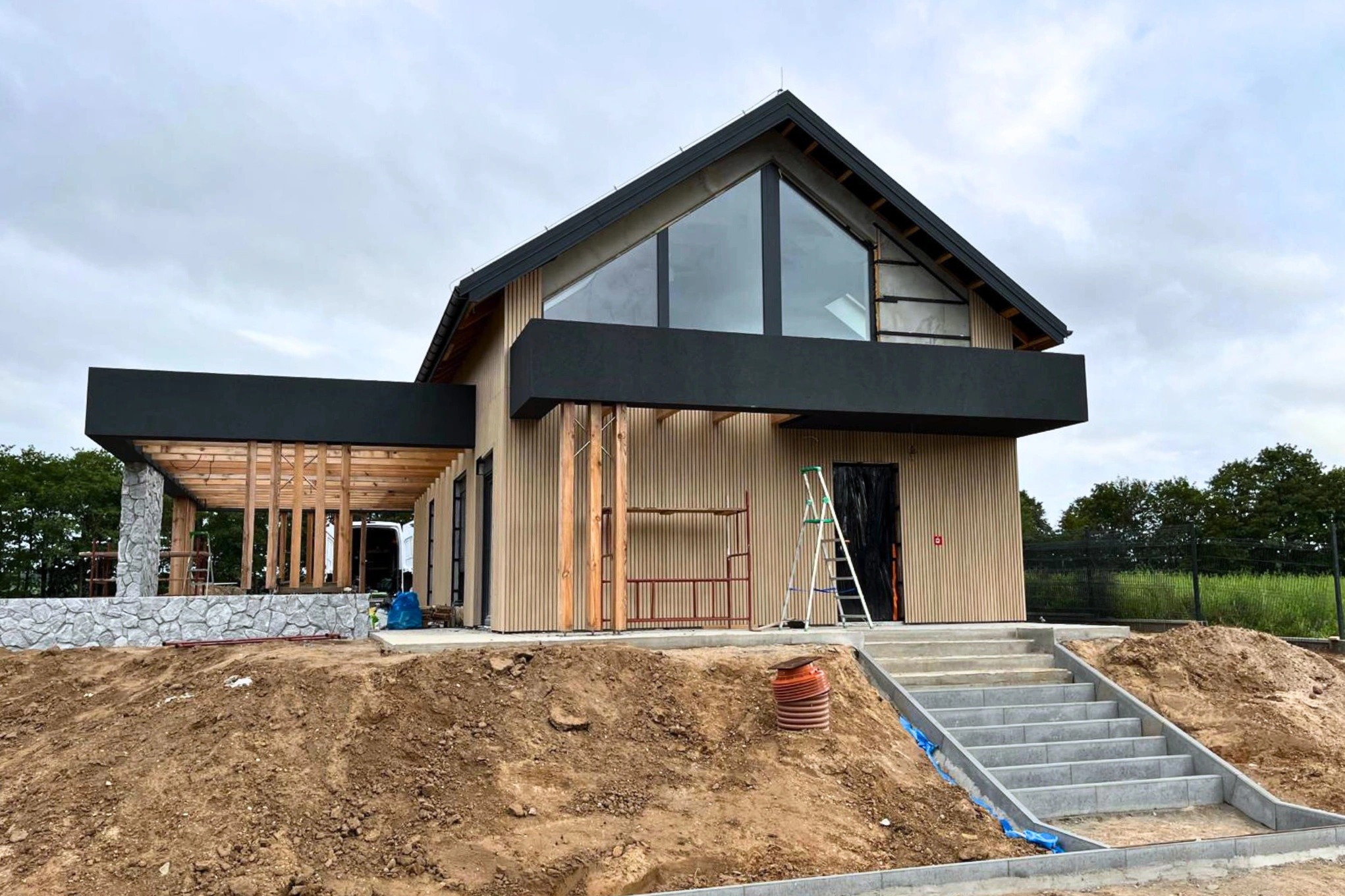 Wznoszony dom jednorodzinny z elewacją z pionowych desek, widoczne rusztowania i schody wejściowe, teren wokół w trakcie prac ziemnych.