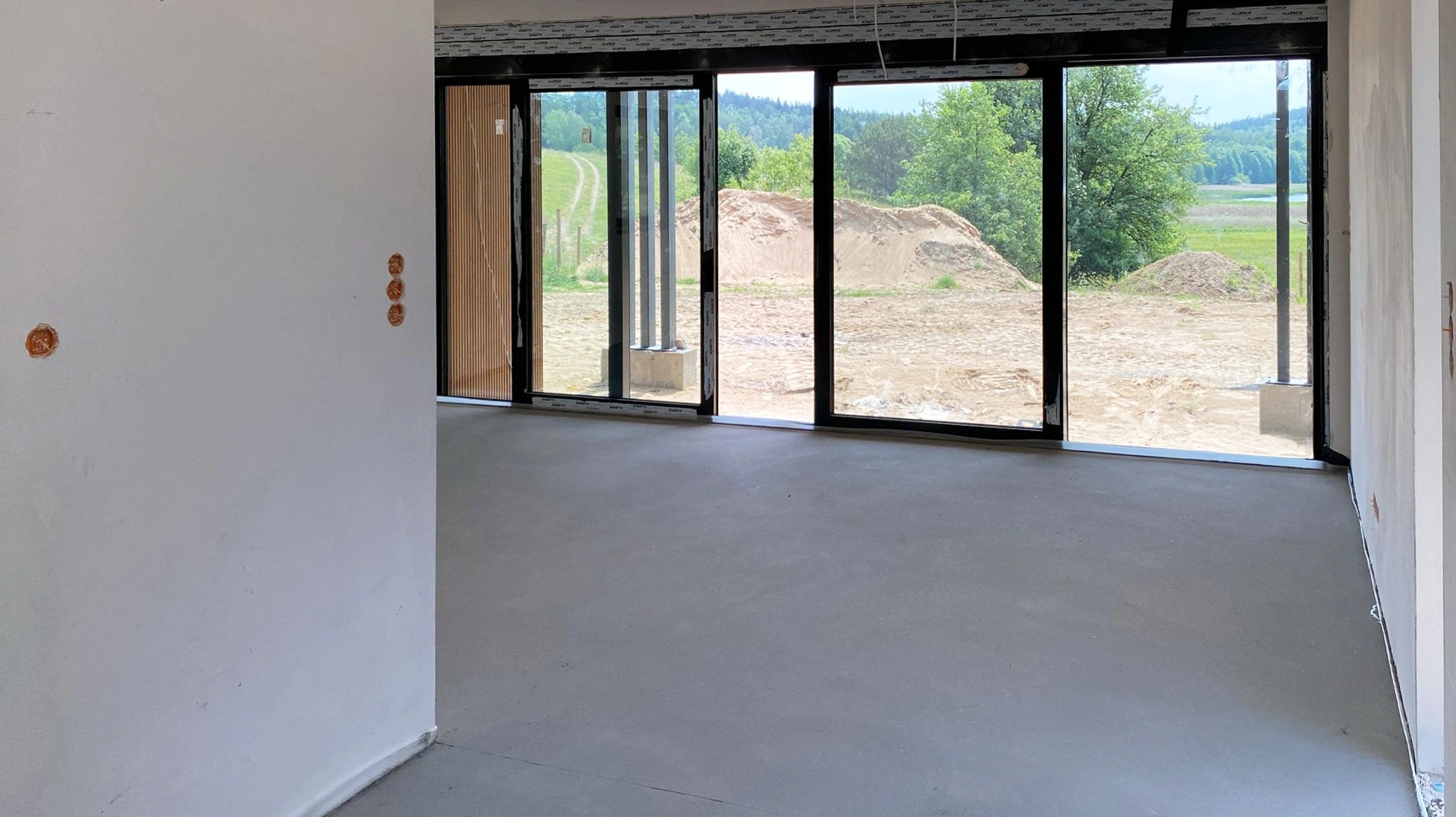 Wnętrze surowego domu z dużymi przesuwnymi oknami z widokiem na pagórkowaty krajobraz i sterty ziemi na zewnątrz, widoczne puszki instalacyjne w ścianie.