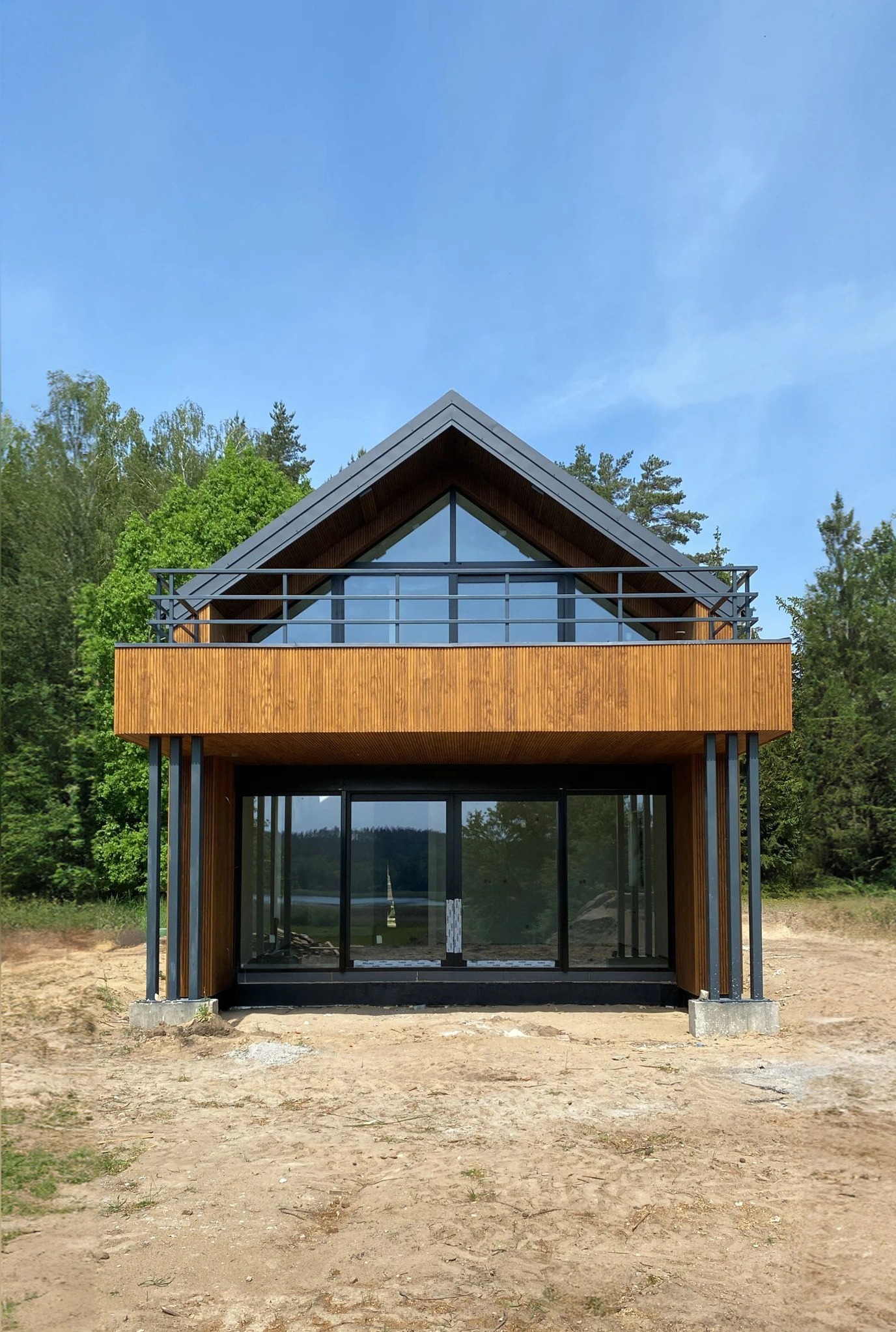Nowoczesny dom z drewnianą elewacją, dużymi przeszkleniami i balkonem z metalową balustradą na tle zieleni, w trakcie budowy.