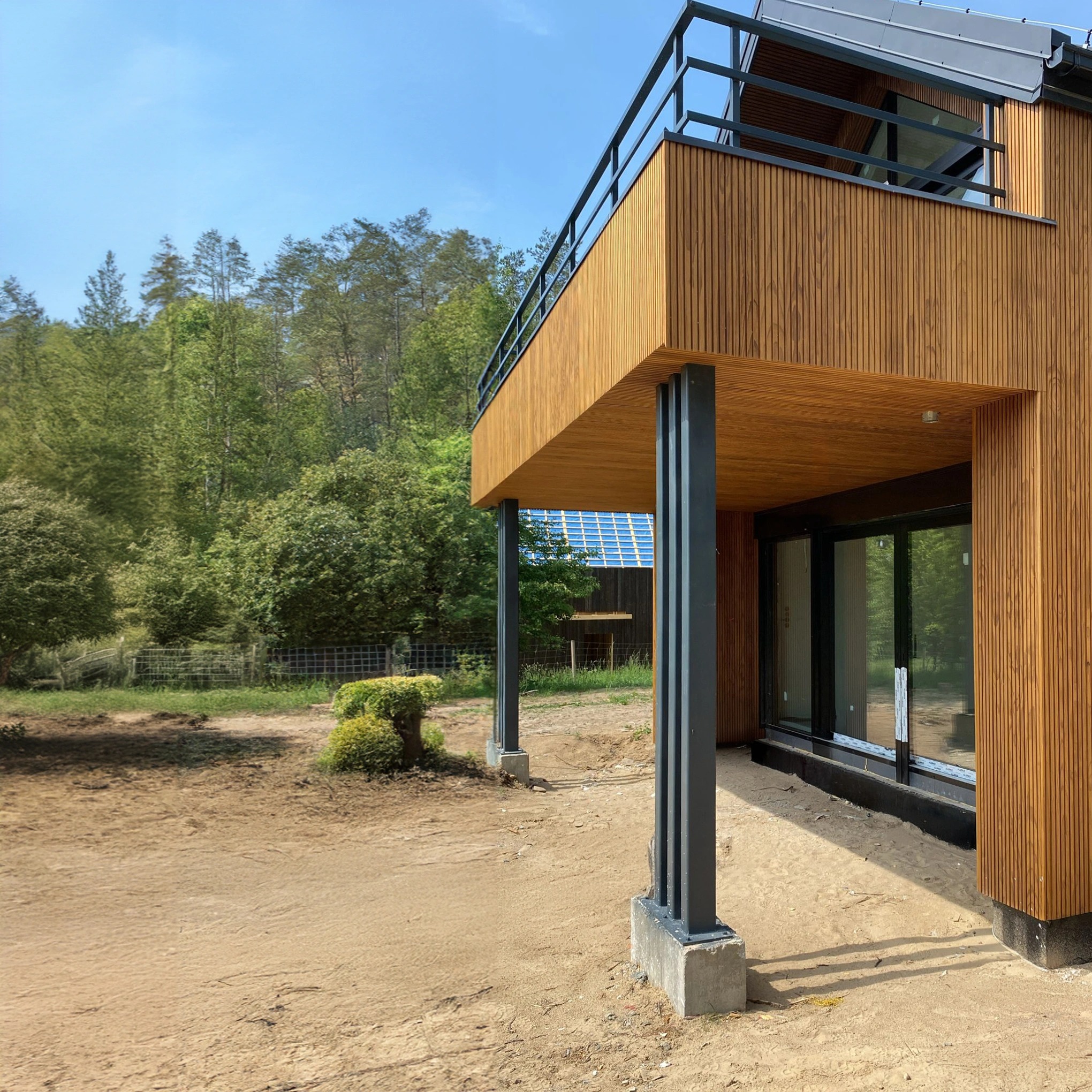Nowoczesny dom z drewnianą elewacją i balkonem, widok z zewnątrz, teren wokół domu w trakcie zagospodarowywania, w tle las i budynek z panelami słonecznymi.