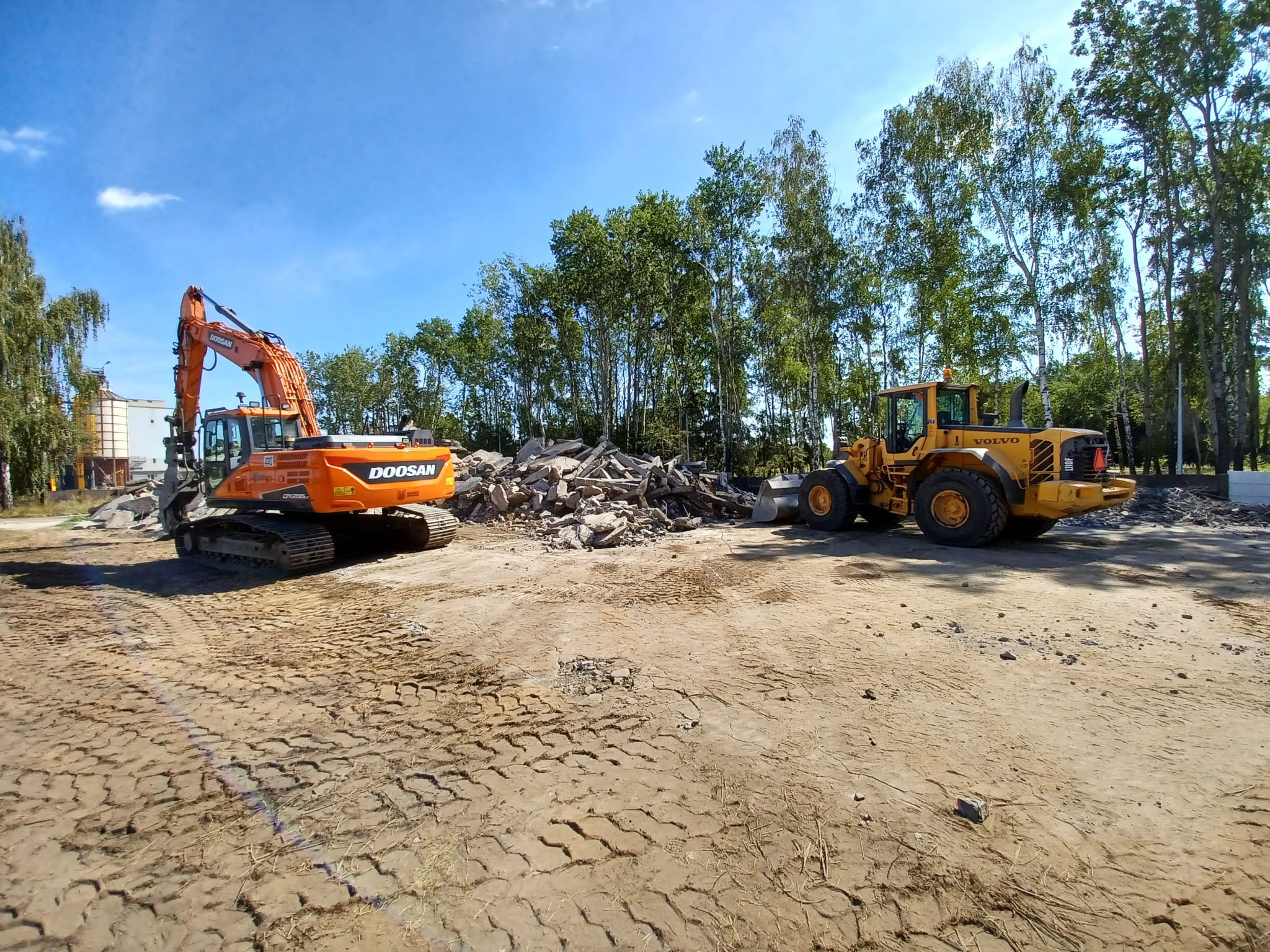 Pomarańczowa koparka Doosan i żółta ładowarka Volvo na placu budowy z gruzem, w tle drzewa i błękitne niebo.