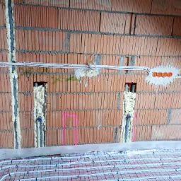 Instalacja ogrzewania podłogowego z widocznymi rurami grzewczymi na folii izolacyjnej, ściana z czerwonej cegły z zamontowanymi puszkami elektrycznymi i przewodami, przestrzenie wypełnione pianką...