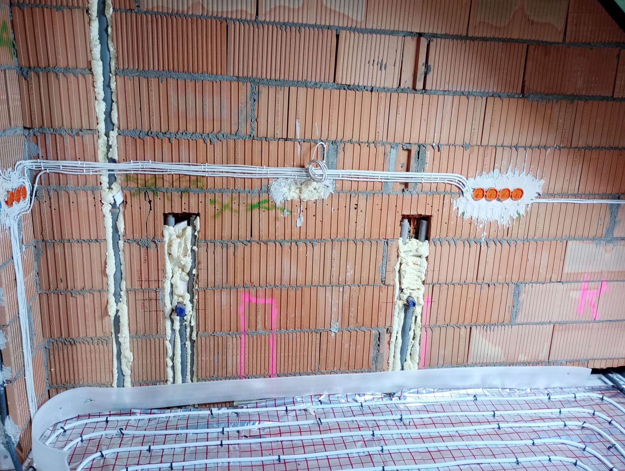 Instalacja ogrzewania podłogowego z widocznymi rurami grzewczymi na folii izolacyjnej, ściana z czerwonej cegły z zamontowanymi puszkami elektrycznymi i przewodami, przestrzenie wypełnione pianką...