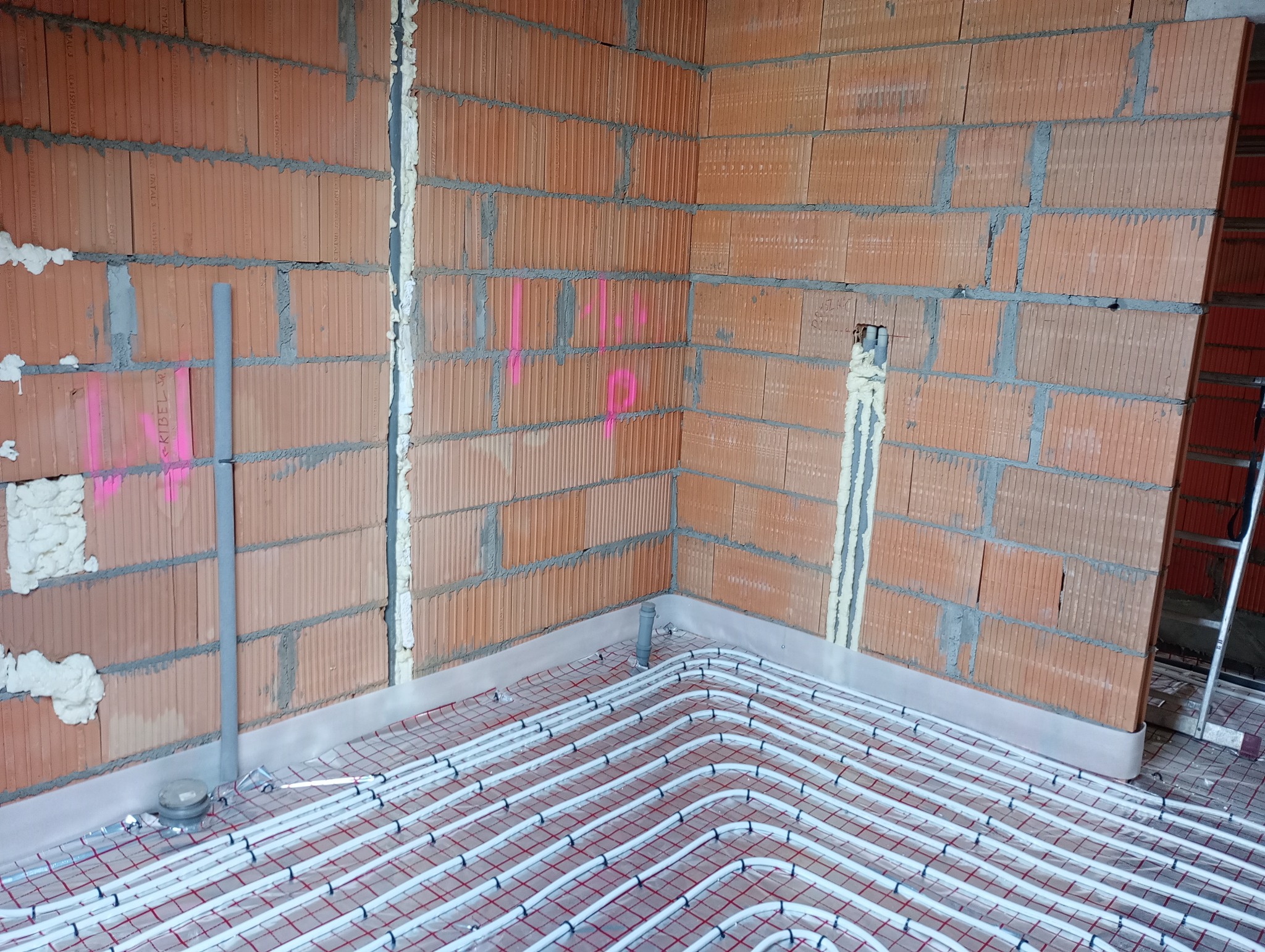 Instalacja ogrzewania podłogowego w surowym wnętrzu z ceglanymi ścianami i pianką montażową w szczelinach.