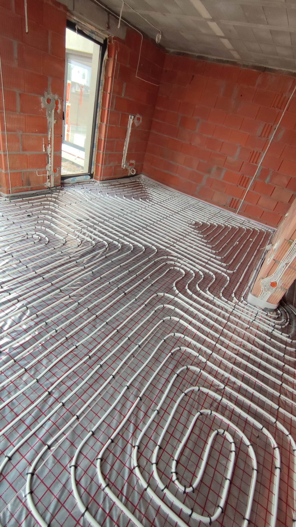 Instalacja ogrzewania podłogowego z białych rur na folii z czerwoną kratką w surowym pomieszczeniu z ceglanymi ścianami i otwartym oknem.