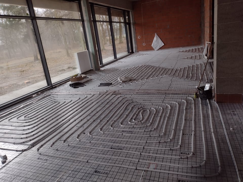 Rozłożone rury ogrzewania podłogowego marki Keller na folii z rastrem w surowym wnętrzu z dużymi oknami i ceglaną ścianą w tle.