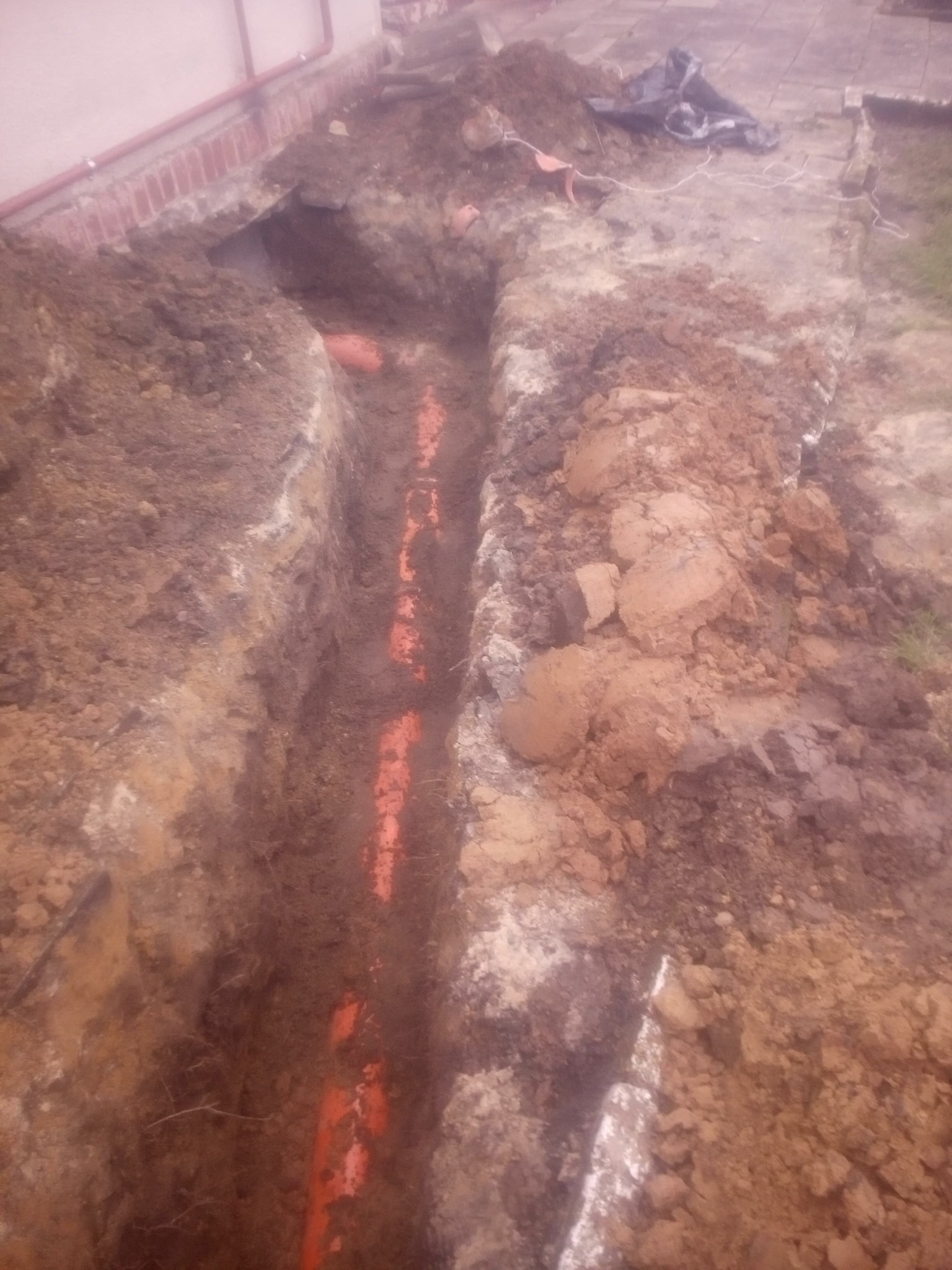 Wykop w ziemi z widocznymi pomarańczowymi rurami instalacyjnymi ułożonymi wzdłuż wykopu, obok fundamentu budynku.