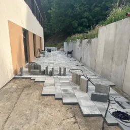 Fragment podwórka z częściowo ułożoną szarą kostką brukową, widoczne przygotowane do ułożenia kostki, czarne kable i ślady butów na piasku.