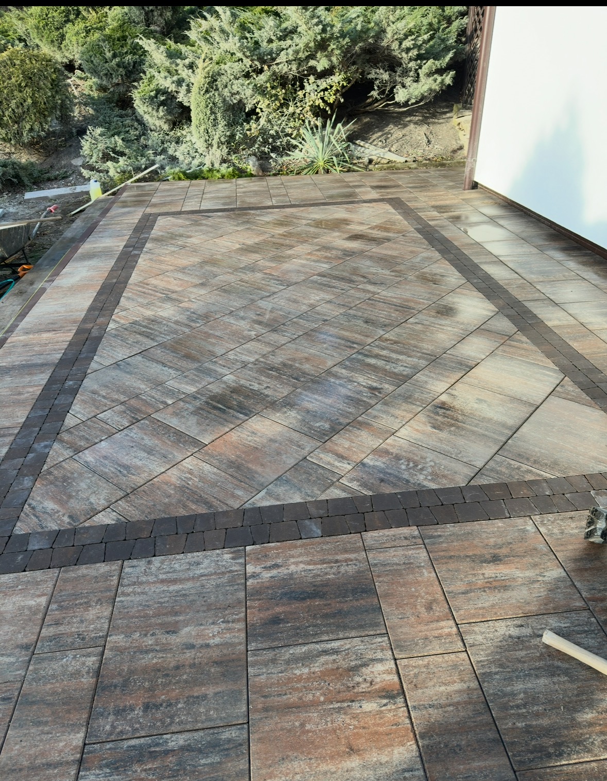 Ułożona kostka brukowa w geometryczny wzór z obramowaniem z ciemniejszej kostki, przylegająca do białej ściany budynku, widoczne narzędzia brukarskie w rogu.