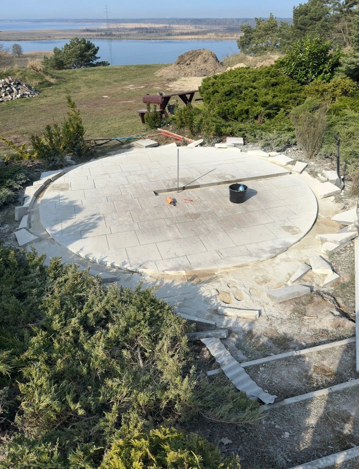 Okazały, okrągły taras z jasnej kostki brukowej w trakcie realizacji, widok z góry, z narzędziami i czarnym wiadrem na powierzchni, niedokończone obrzeże z kostki, w tle malowniczy krajobraz...