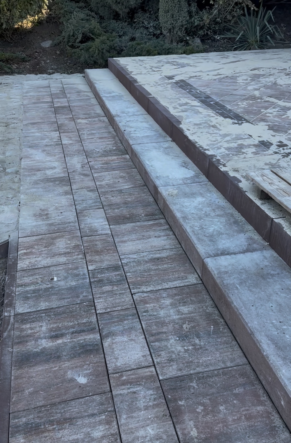 Ułożona ścieżka z kostki brukowej w odcieniach szarości i brązu, z widocznym obrzeżem i fragmentem powierzchni przygotowanej pod dalsze prace, tło z zielenią.