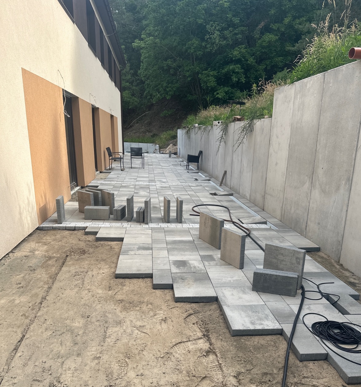 Fragment podwórka z częściowo ułożoną szarą kostką brukową, widoczne przygotowane do ułożenia kostki, czarne kable i ślady butów na piasku.