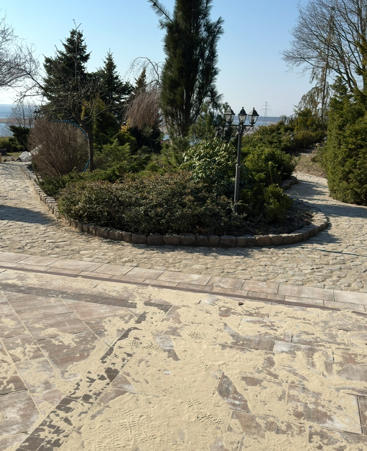 Ścieżka z kostki brukowej, częściowo zasypana piaskiem, prowadząca do okrągłego klombu z różnorodną roślinnością, w tle jezioro i słupy wysokiego napięcia.