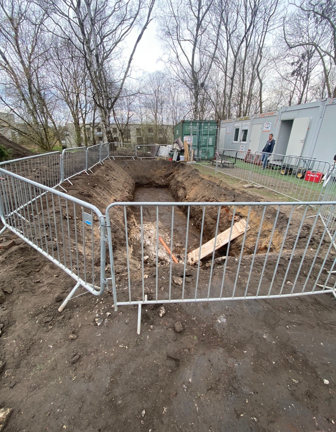 Wykop zabezpieczony metalowym ogrodzeniem tymczasowym, widoczne ślady prac ziemnych, w tle kontener i barak budowlany, drzewa bezlistne.