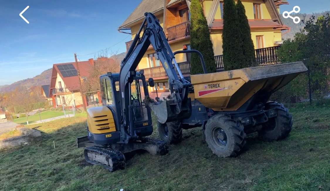 Żółto-czarny mini-koparka Volvo nabiera ziemię do żółtej wywrotki Terex na trawiastym terenie z domami w tle.