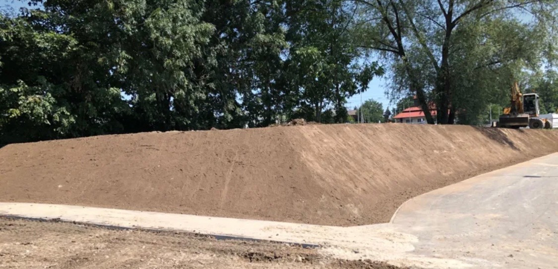 Świeżo usypany nasyp ziemny z równą powierzchnią, widoczna krawędź betonowa i koparka w tle, otoczona zielenią drzew.