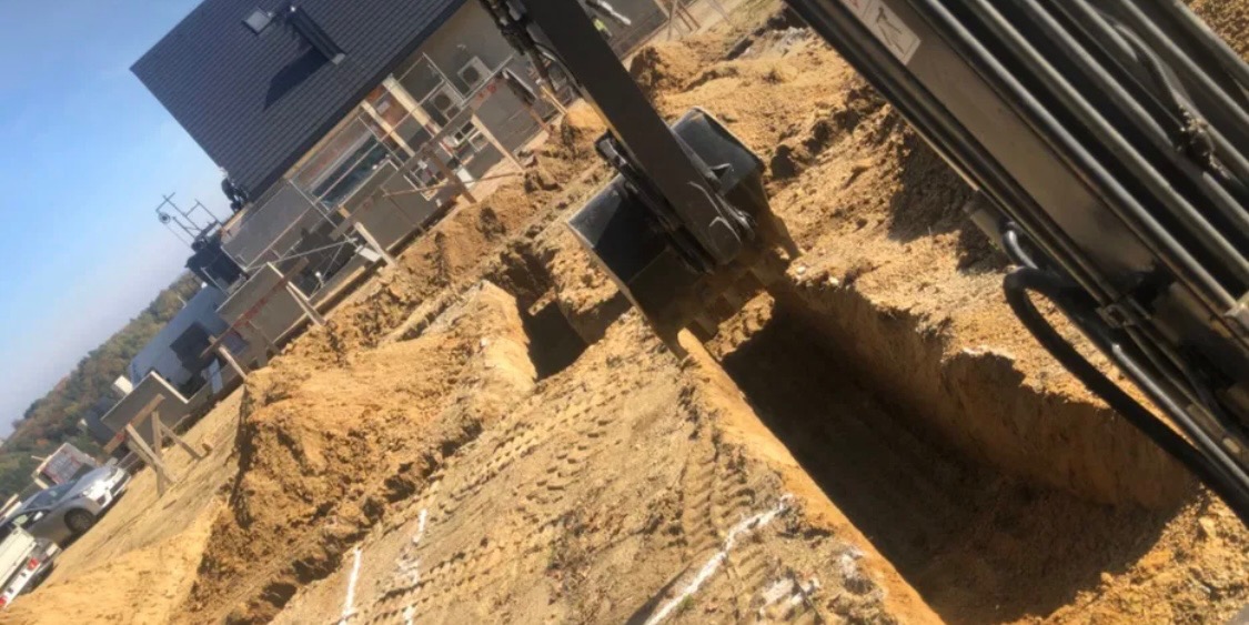 Koparka zagłębia się w ziemię, tworząc wykop pod fundamenty nowego domu, widoczny w tle pod rusztowaniem.