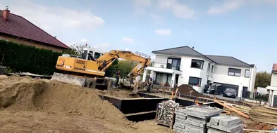Żółta koparka przy wykopie fundamentów pod budynek w Tymbarku, widoczne szalunki i ekipa budowlana.