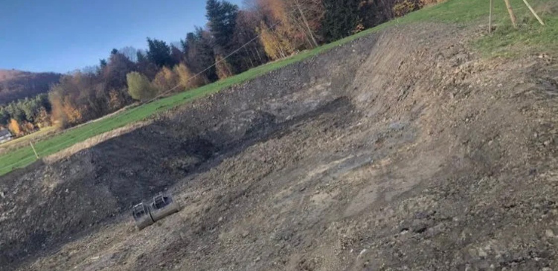 Skarpa ziemna po wykopie, widoczne rury betonowe w dolnej części zbocza, w tle drzewa i fragment zielonego terenu.
